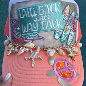 May include: A coral-colored trucker hat featuring a rectangular patch with the text LAID BACK Since WAY BACK, three surfboard appliques, a gold chain draped across the brim, and charms including seashells, white pearls, and gold starfish.