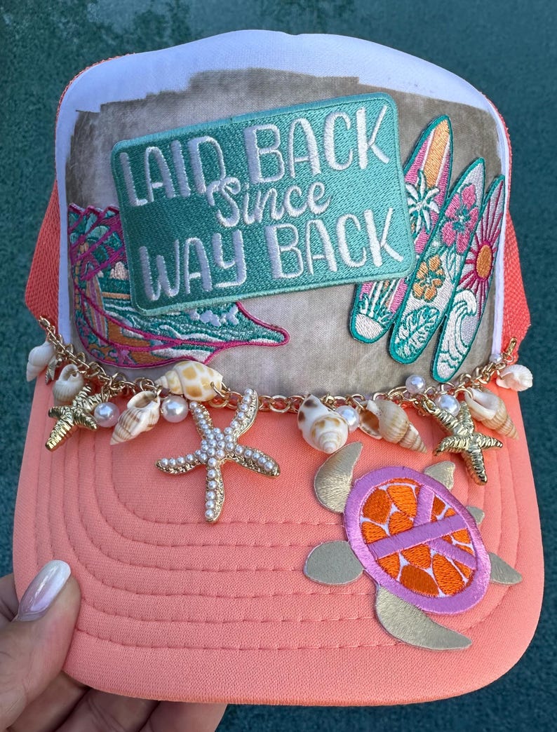 May include: A coral-colored trucker hat featuring a rectangular patch with the text LAID BACK Since WAY BACK, three surfboard appliques, a gold chain draped across the brim, and charms including seashells, white pearls, and gold starfish.