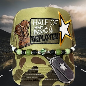 May include: Camouflage trucker hat with the text "HALF OF MY heart is DEPLOYED". The hat features a patch with brown boots, a star, and a dog tag pendant with a star. A beaded accent is attached to the brim.