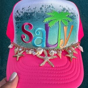 May include: A bright neon pink trucker hat featuring a white front panel with a teal and white ocean wave print. The word Salty is printed in colorful letters with a green palm tree. A gold chain with small seashells and starfish accents.