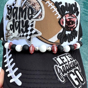 May include: A white and black trucker hat featuring football-themed patches with the text "game day", "touch down", and "LET'S freaking GO". The hat is decorated with a beaded trim of black, white, and football-shaped beads across the brim.