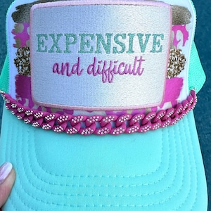 May include: A turquoise trucker hat with a white mesh back and a pink chain detail. The hat features a rectangular patch with the words "EXPENSIVE and difficult" embroidered in green and pink.