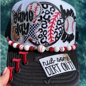 May include: Black and white baseball cap with a white front panel featuring a "Game Day" design, a baseball, and a leopard print baseball. The brim has red "T" shapes and a patch that says "rub some dirt on it". A beaded necklace with baseballs and red beads is attached.