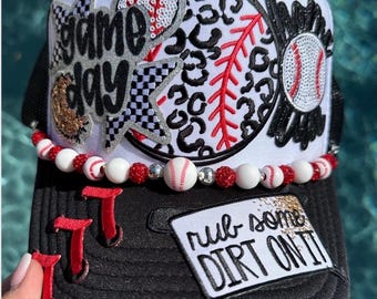 May include: Black and white baseball cap with a white front panel featuring a "Game Day" design, a baseball, and a leopard print baseball. The brim has red "T" shapes and a patch that says "rub some dirt on it". A beaded necklace with baseballs and red beads is attached.