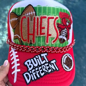 May include: A red and white trucker hat featuring a green football field graphic, a glittery gold and red CHIEFS patch, a red rhinestone chain, and a white patch with the black text BUILT DIFFERENT, alongside football and bow appliques.