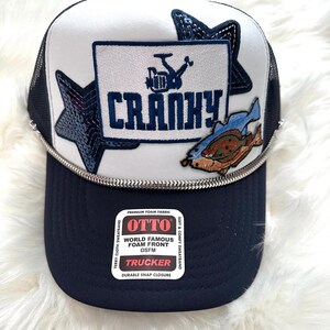 Op de afbeelding: Een witte en blauwe trucker-pet met een mesh-rug. De pet heeft een patch met de tekst "CRANKY" met een cartoon-afbeelding van een vrachtwagen. De pet heeft ook een blauwe ster en een vis-patch.
