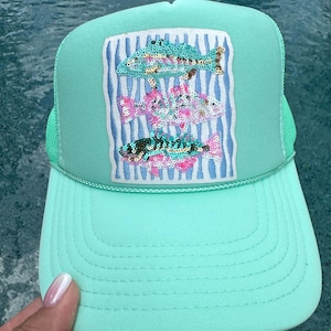 May include: A mint green trucker hat featuring a rectangular patch on the front. The patch displays three stacked fish designs created with colorful sequins against a background of vertical blue and white wavy stripes. The hat has a mesh back.