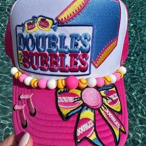 May include: A bright pink and white trucker hat featuring an embroidered patch that reads "DOUBLES & BUBBLES" in blue and pink lettering. The hat is decorated with a row of pink, white, and yellow beads and a large, colorful bow patch on the brim.