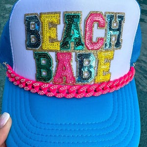 May include: A bright blue trucker hat with a white front panel featuring the text BEACH BABE in multi-colored sequined letters with gold glitter outlines. A vibrant pink plastic chain with embedded rhinestones accents the base of the front panel.