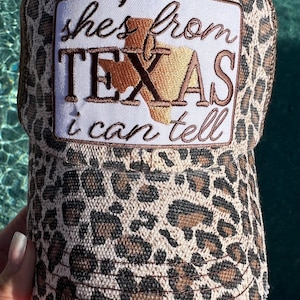 May include: A leopard print baseball cap with a rectangular white patch on the front. The patch features the embroidered text 'she's from TEXAS i can tell' in brown, with a gold-colored embroidered outline of the state of Texas in the center.