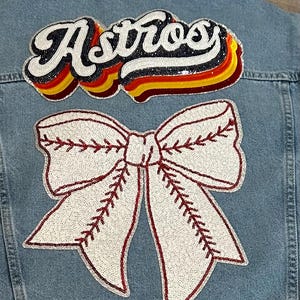 May include: A denim jacket with a white and red sequined bow and a sequined "Astros" patch in rainbow colors.