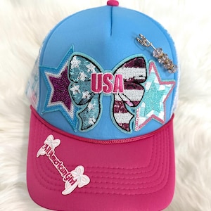 Op de afbeelding: Een blauw-roze truckerpet met een strik- en sterontwerp. De strik heeft het woord "USA" en is versierd met pailletten. De pet heeft ook de tekst "All American Girl" op de rand. Een decoratieve speld met kralen is aan de zijkant bevestigd.