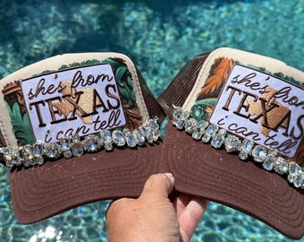 May include: Two brown trucker hats featuring a white rectangular patch with the text "she's from TEXAS i can tell" and a tan Texas state outline. The base of each patch is adorned with a row of clear, faceted rhinestones across the brim.