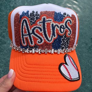 May include: Orange trucker hat with the word "Astros" in sparkling letters. The hat features a baseball heart patch and a rhinestone band. The top panel has a mix of patterns in orange, blue, and leopard print.