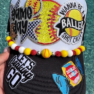 May include: Black and white baseball cap with patches and text. The cap features a sequined softball design, the words "Game Day", "Wanna Be A Baller Shot Caller", and "Let's Freaking Go". A beaded necklace with a softball charm is attached.