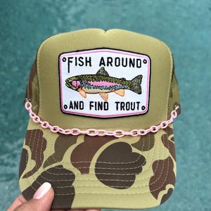 May include: A camouflage trucker hat featuring a rectangular patch with a colorful trout illustration and the text FISH AROUND AND FIND TROUT. The hat is accented with a light pink plastic chain draped across the front above the brim.