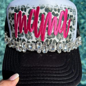 May include: A trucker hat featuring a white front panel with a gray leopard print pattern and the word mama in bright pink script. A row of clear, faceted rhinestones lines the base of the front panel, and the brim and back are black.