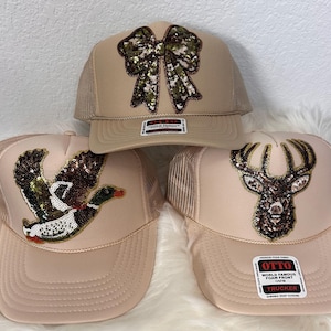 May include: Three beige trucker hats with mesh backs and foam fronts. The top hat features a sequined camouflage bow. The bottom left hat displays a sequined mallard duck in flight. The bottom right hat shows a sequined deer head silhouette.