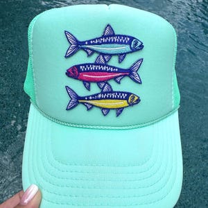 May include: A mint green trucker hat featuring an embroidered patch of three stacked fish. The top fish is blue, the middle fish is pink with a purple head, and the bottom fish is yellow with a blue head. The hat has a mesh back and a solid front.