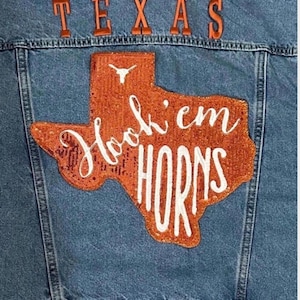 May include: A blue denim jacket featuring the word TEXAS in orange letters across the top. Below, a large orange sequined patch shaped like the state of Texas displays a white longhorn logo and the white embroidered text Hook 'em HORNS.