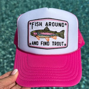 May include: A bright pink and white trucker hat featuring a rectangular patch on the front. The patch displays an embroidered trout fish with colorful details and the black text "FISH AROUND AND FIND TROUT" with small black circles at the corners.