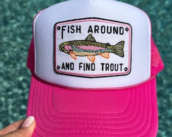 May include: A bright pink and white trucker hat featuring a rectangular patch on the front. The patch displays an embroidered trout fish with colorful details and the black text "FISH AROUND AND FIND TROUT" with small black circles at the corners.