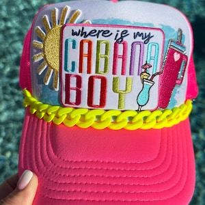 May include: A bright pink trucker hat featuring a white front panel with a patch that reads "where is my CABANA BOY" in colorful letters. The patch includes a yellow sun, a blue cocktail, and a pink tumbler. A neon yellow chain trim accents the brim.