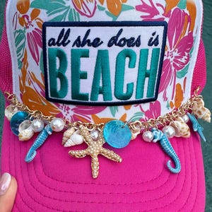 May include: A pink trucker hat with a white front panel featuring a floral design and the text "all she does is BEACH." The hat is adorned with a gold chain and seashell charms, including a starfish, seahorses, and pearls.