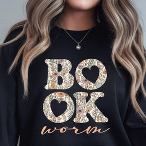 May include: Black sweatshirt with the words "BOOK" and "OK" in large, floral-filled letters, with heart-shaped cutouts. The word "worm" is written in a cursive font below. The sweatshirt is a comfortable, casual style.