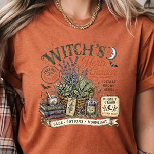 May include: A burnt orange t-shirt featuring a graphic print of a basket filled with herbs, a mortar and pestle on stacked books, and a bottle labeled Witches Brew. The text reads Witch's Herb Garden, Freshly Gathered, Locally Grown Herbs, Moonlit Grown, Autumn Picked, Sage, Potions, and Moonlight.