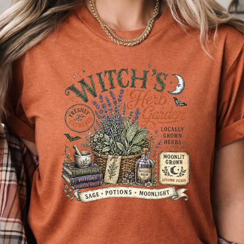 May include: A burnt orange t-shirt featuring a graphic print of a basket filled with herbs, a mortar and pestle on stacked books, and a bottle labeled Witches Brew. The text reads Witch's Herb Garden, Freshly Gathered, Locally Grown Herbs, Moonlit Grown, Autumn Picked, Sage, Potions, and Moonlight.