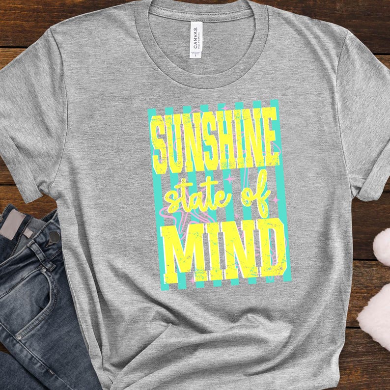 K&ouml;nnte beinhalten: Graues T-Shirt mit dem Text "SUNSHINE state of MIND" in Gelb und T&uuml;rkis. Der Text ist in einem Blockdesign mit einer Sternengrafik gehalten. Das Shirt wird mit einer blauen Jeans kombiniert.