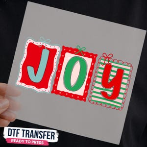 May include: A DTF transfer sheet featuring the word JOY in three separate rectangular frames. The J is light blue on a red background, the O is green on a cream background, and the Y is red on a green and white striped background.
