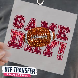 May include: A DTF transfer sheet featuring the text GAME DAY! in a red glitter finish with a white outline. Centered over the text is a graphic of an American football with a textured orange glitter surface and silver glitter laces and stripes.