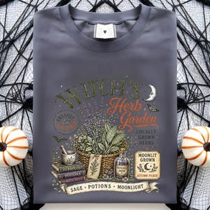 May include: A dark gray crew neck t-shirt featuring a graphic print of a basket of herbs, a mortar and pestle, books, and a bottle labeled Witches Brew. The text reads Witch's Herb Garden, Freshly Gathered, Locally Grown Herbs, and Moonlit Grown.