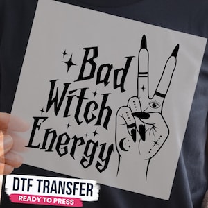 May include: A black graphic design on a clear film featuring the text Bad Witch Energy in a stylized font, accompanied by small star icons and an illustration of a hand making a peace sign with an eye, moon, and dot details on the fingers.