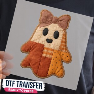 May include: A DTF transfer featuring a ghost-shaped design with a patchwork aesthetic in shades of cream, orange, and brown. The ghost has a brown bow on its head, two black oval eyes, and a small black mouth, all outlined in a textured gold border.