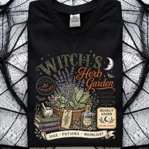 May include: A black t-shirt featuring a graphic print with the text Witch's Herb Garden. The design includes a woven basket of herbs, a mortar and pestle, stacked books labeled Herbology, Potions, and Plants & Spells, and a bottle of Witches Brew.