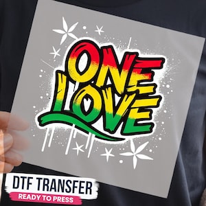 May include: A square DTF transfer sheet featuring the text ONE LOVE in a bold, graffiti-style font. The letters transition from red at the top to yellow in the middle and green at the bottom. White star-like accents and paint splatters surround the text.