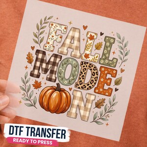 May include: A DTF transfer sheet featuring the text FALL MODE ON in block letters with various patterns including plaid, leopard print, and floral designs. An orange pumpkin sits below the text, surrounded by small leaves, vines, and hearts.