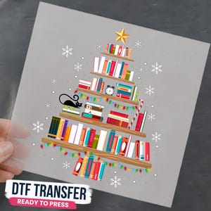 May include: A DTF transfer sheet featuring a graphic of a Christmas tree constructed from stacked colorful books on wooden shelves. The design includes a yellow star topper, a black cat, a candy cane, string lights, and white snowflakes on a gray background.