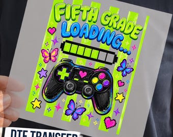May include: A DTF transfer sheet featuring the text FIFTH GRADE LOADING in blue and green, a black video game controller, a loading bar, colorful butterflies, pixelated hearts, and stars against a background of vertical lime green stripes.