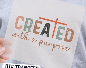 May include: A square DTF transfer sheet featuring the text "CREATED with a purpose" in a multi-colored, distressed font. The word "CREATED" is in large block letters with a cross replacing the letter "T", while "with a purpose" is in script.