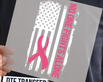 May include: A DTF transfer sheet featuring a distressed American flag design in white and gray. A bright pink awareness ribbon is positioned over the stripes. The text NO ONE FIGHTS ALONE is printed vertically in pink capital letters next to the flag.