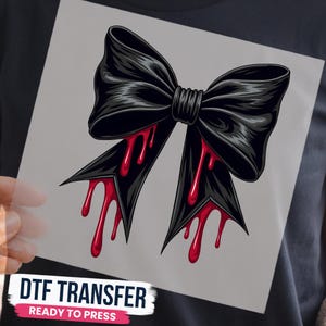 May include: A black bow graphic with red liquid dripping from the bottom edges. The design is printed on a square transfer sheet with the text DTF TRANSFER READY TO PRESS visible at the bottom of the image against a dark fabric background.