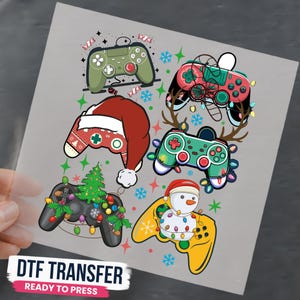 May include: A DTF transfer sheet featuring six video game controllers decorated with holiday themes, including a Santa hat, a snowman, a Christmas tree, reindeer antlers, and colorful string lights, accented by snowflakes and candy motifs.