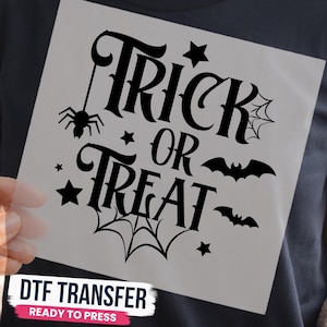 May include: A black DTF transfer sheet featuring the text Trick or Treat in a decorative font. The design includes a spider hanging from a web, two flying bats, and several stars. The graphic is printed on a clear film for heat application.