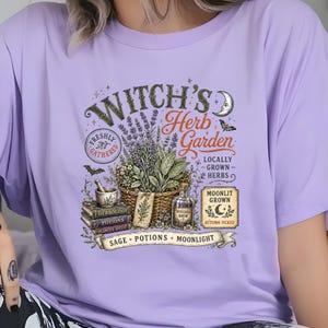 May include: A light purple t-shirt featuring a graphic print with the text "Witch's Herb Garden". The design includes a basket of herbs, a mortar and pestle on stacked books, a glass bottle, a crescent moon, and the words "Sage, Potions, Moonlight".