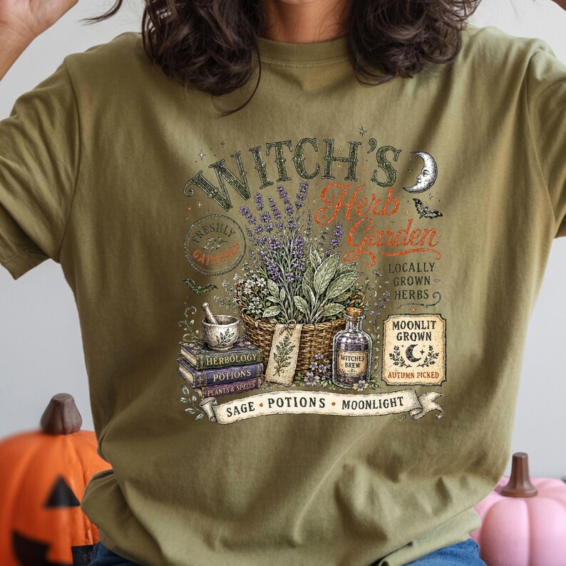 May include: An olive green t-shirt featuring a graphic print with the text Witch's Herb Garden, Moonlit Grown, and Sage Potions Moonlight. The design includes a basket of lavender, a mortar and pestle, stacked books, and a potion bottle.