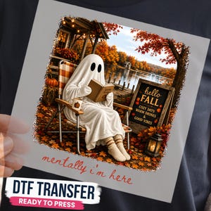 May include: A DTF transfer featuring a ghost in a white sheet sitting in a folding chair reading a book by a lake with autumn trees. A sign reads hello FALL COZY DAYS WARM DRINKS & GOOD VIBES. The text mentally i'm here is printed below.